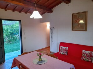 Um quarto com uma mesa posta para refeições, um sofá vermelho com almofadas florais, um lustre pendurado e uma porta que dá para um jardim.