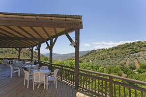 Um deck de madeira com cadeiras e mesas brancas, com vista para um vinhedo e colinas.