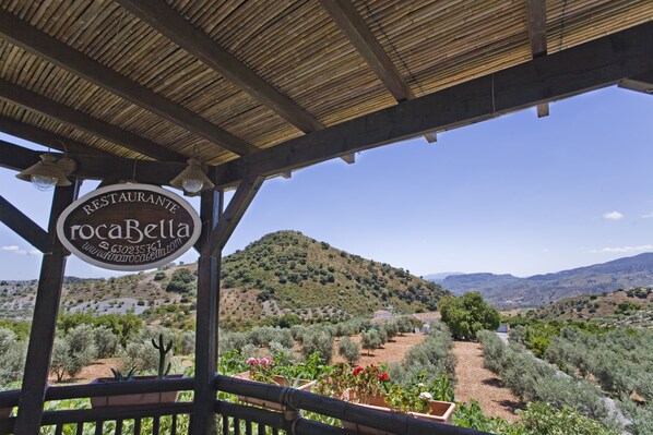 Placa de restaurante com o nome 'RocaBella' e um endereço de site, com paisagem montanhosa ao fundo, incluindo cactos e arbustos.