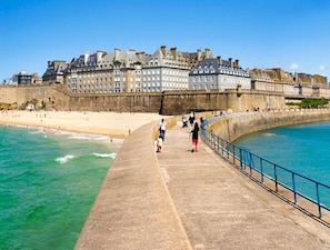 A coastal promenade with a stone walkway, a beach, and a historic building with multiple stories and a fortified wall. - Great times with family and friends (Mont-Dol)