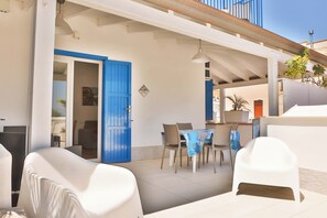 Un espace extérieur abrité, aménagé pour prendre les repas, avec une table et des chaises, une porte bleue et un mur blanc.