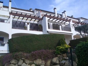 A white building with a balcony, a stone wall, and a hedge.