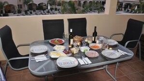 A dining table set for two with plates, glasses, and bottles, surrounded by chairs, on a balcony with a view of hotel buildings and palm trees. - 2 Bed Luxury Apartment Overlooking The Lush Gardens And Pools. With Spa And Gym For Guests (Mandria)