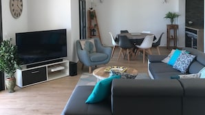 A modern living room with a large flat-screen TV, a grey sofa with teal cushions, a dining area with a table and chairs, and a wooden ceiling.