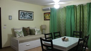 A small, cozy room with a bed, a dining table, and chairs. There is a ceiling fan, a wall-mounted air conditioner, and a framed picture on the wall.