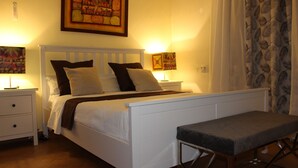 A bedroom with a white bed, two bedside tables, a bench, and a large painting on the wall.