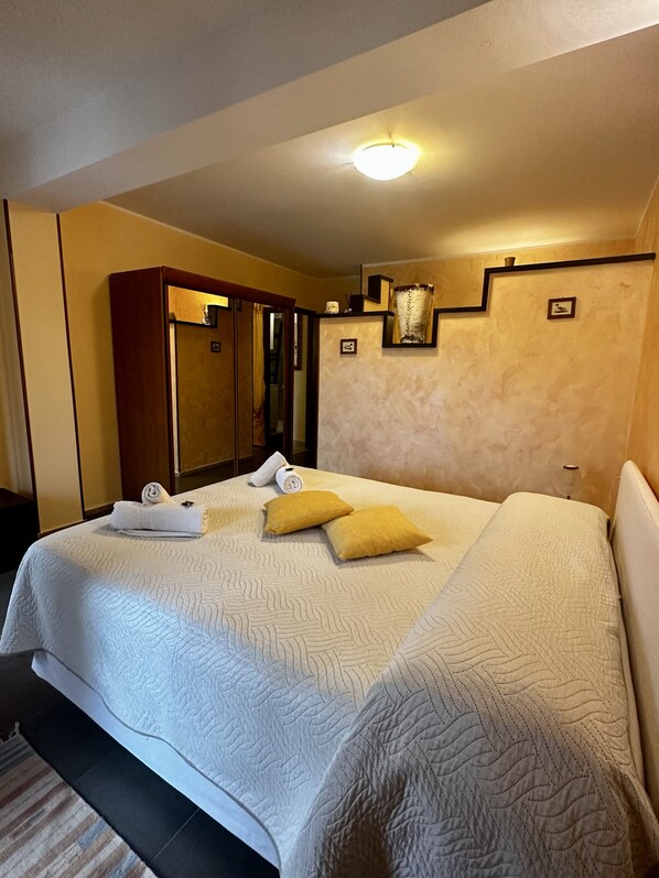 Une chambre d’hôtel avec un grand lit, une armoire, un miroir et une petite table avec une lampe.