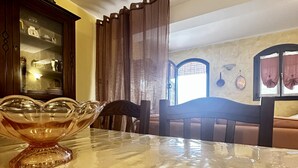Una mesa de comedor con un centro de mesa de cristal, un aparador de madera con puertas de vidrio y una cama con cabecera en una habitación con pared de piedra y una ventana con cortinas.