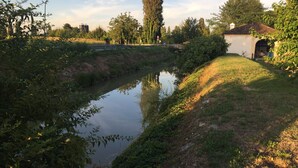 Escena tranquila junto al río, con una orilla cubierta de hierba, un puente y un edificio con una entrada en arco.