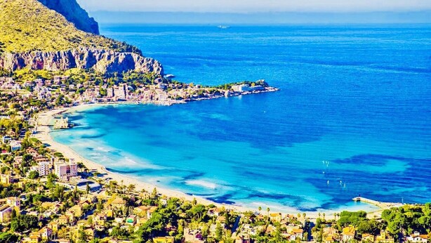 A coastal town with a beach, clear blue waters, and a mountainous backdrop.