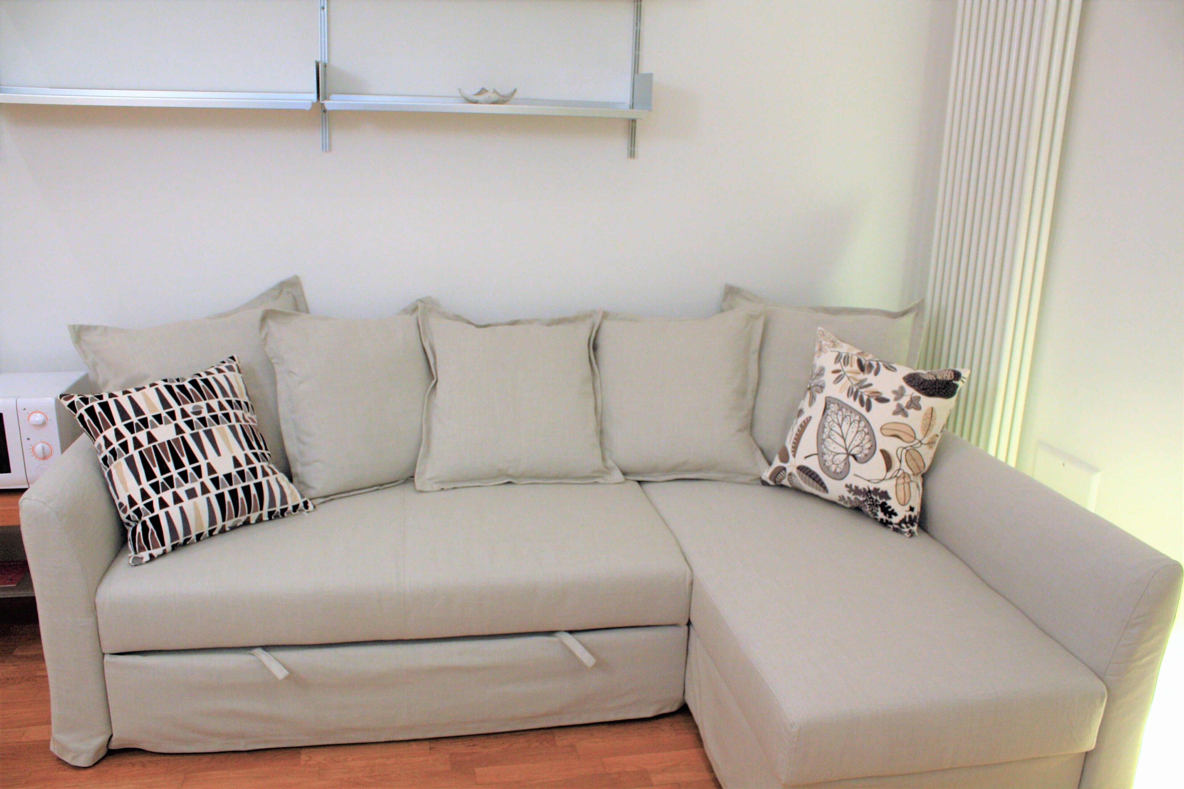 A beige sectional sofa with multiple cushions, a shelf above, and a window with blinds.