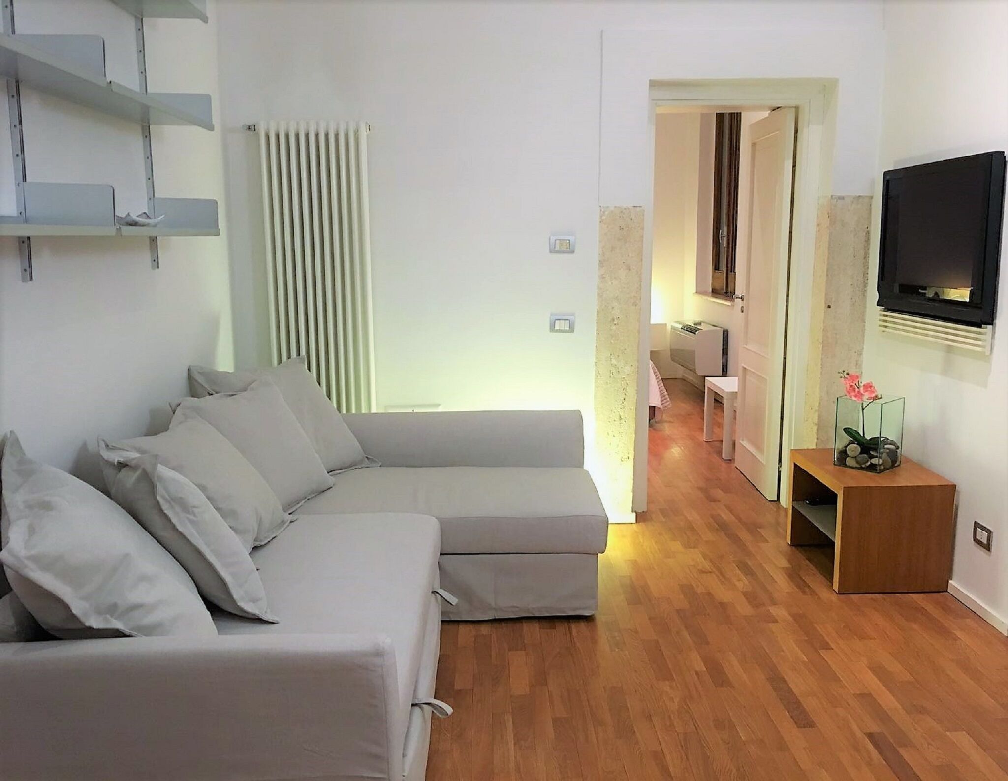 A modern living room with a grey sectional sofa, wooden flooring, a built-in TV, and a small wooden side table with a vase.