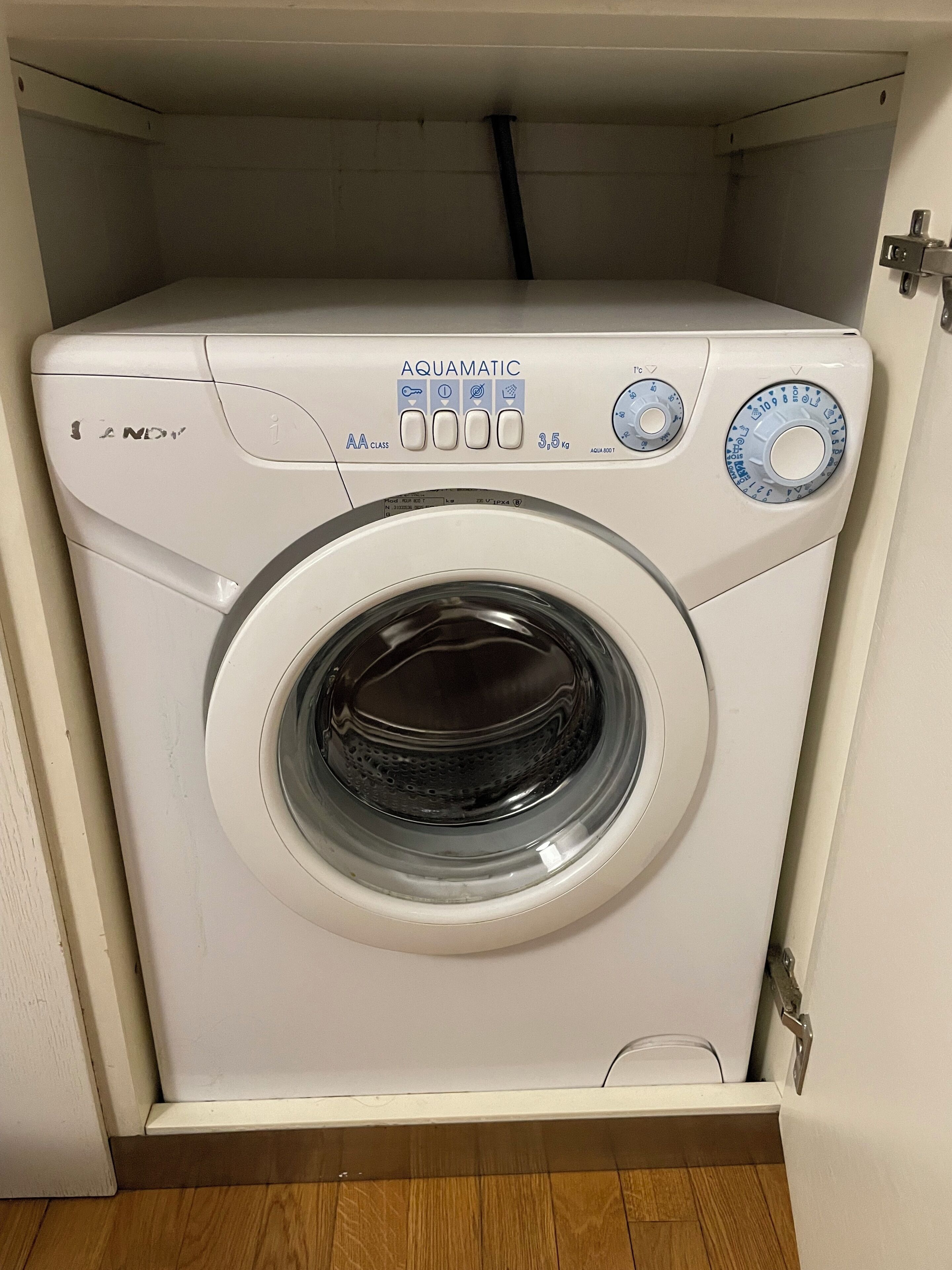 A front-loading white washing machine with a control panel, located in a laundry room with wooden flooring.