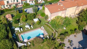 Vista aérea de un vecindario con piscina rodeada de sillones de descanso y sombrillas, un jardín y casas colindantes.