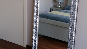 Ein Zimmer mit einem großen Spiegel mit strukturierter Rahmen, einem Bett mit blauer Bettdecke, einem Nachttisch mit Lampe und einer Tür mit Griff.