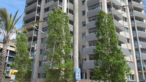 Ein hoch aufragendes Wohngebäude mit mehreren Balkonen, einem Fußgängerübergang und einer Straße gesäumt von Palmen.