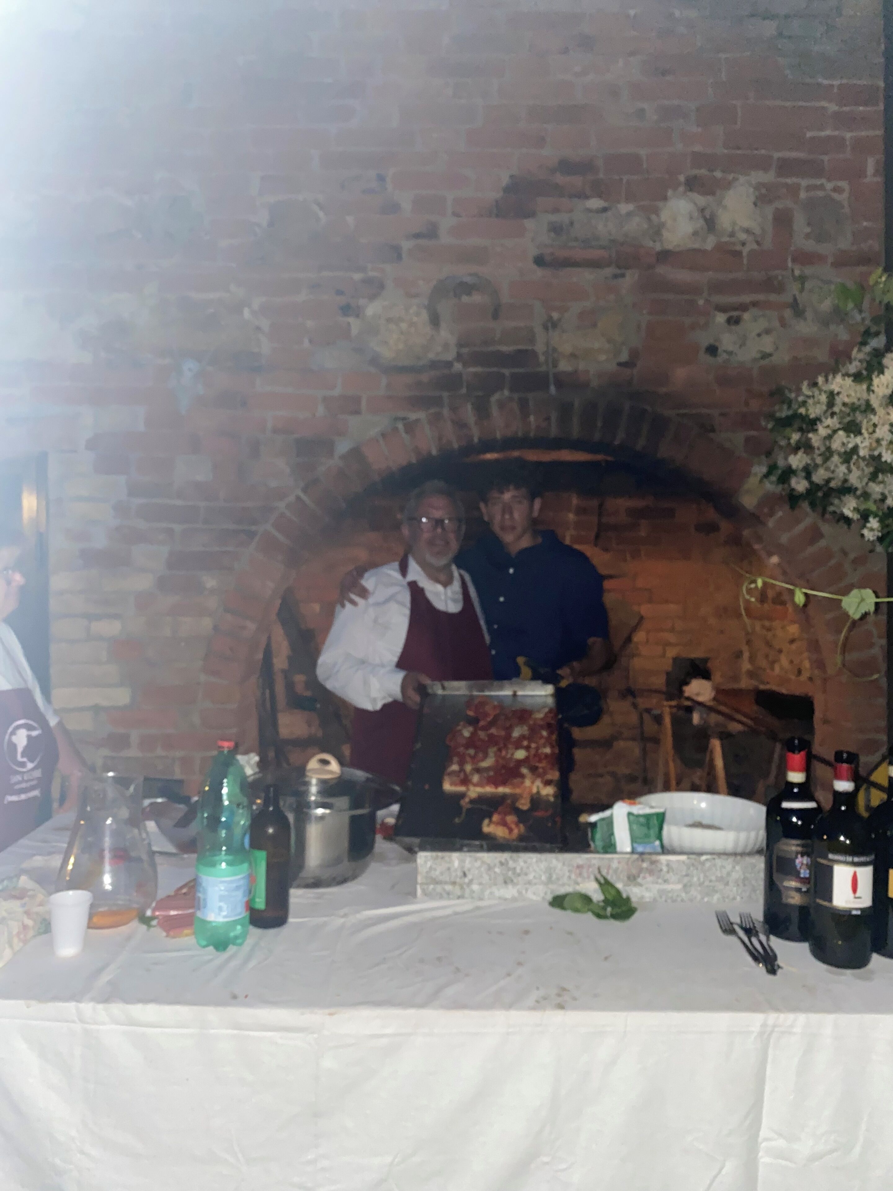 A brick oven with pizza being cooked, a table with bottles and a green bottle, and a person in a white apron.