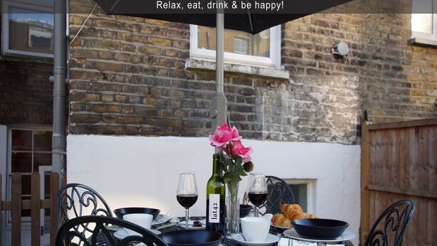 An outdoor dining table with a bottle of wine, glasses, a bowl, and a croissant. - Brilliant Fulham Experience - Garden (Private) Apartment (London)