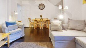 A modern living room with a sofa, a smaller couch, a dining table with chairs, a clock on the wall, and a lamp. - Brilliant Fulham Experience - Garden (Private) Apartment (London)