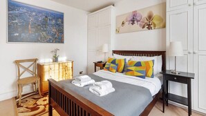 A neatly arranged bedroom with a cityscape painting, a floral artwork, a wooden dresser, and a bed with colorful pillows. - Brilliant Fulham Experience - Garden (Private) Apartment (London)
