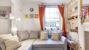 A cozy living room with a grey sofa, multiple pillows, a side table, a lamp, a window with orange curtains, and a bookshelf with plants. - Brilliant Fulham Experience - Garden (Private) Apartment (London)