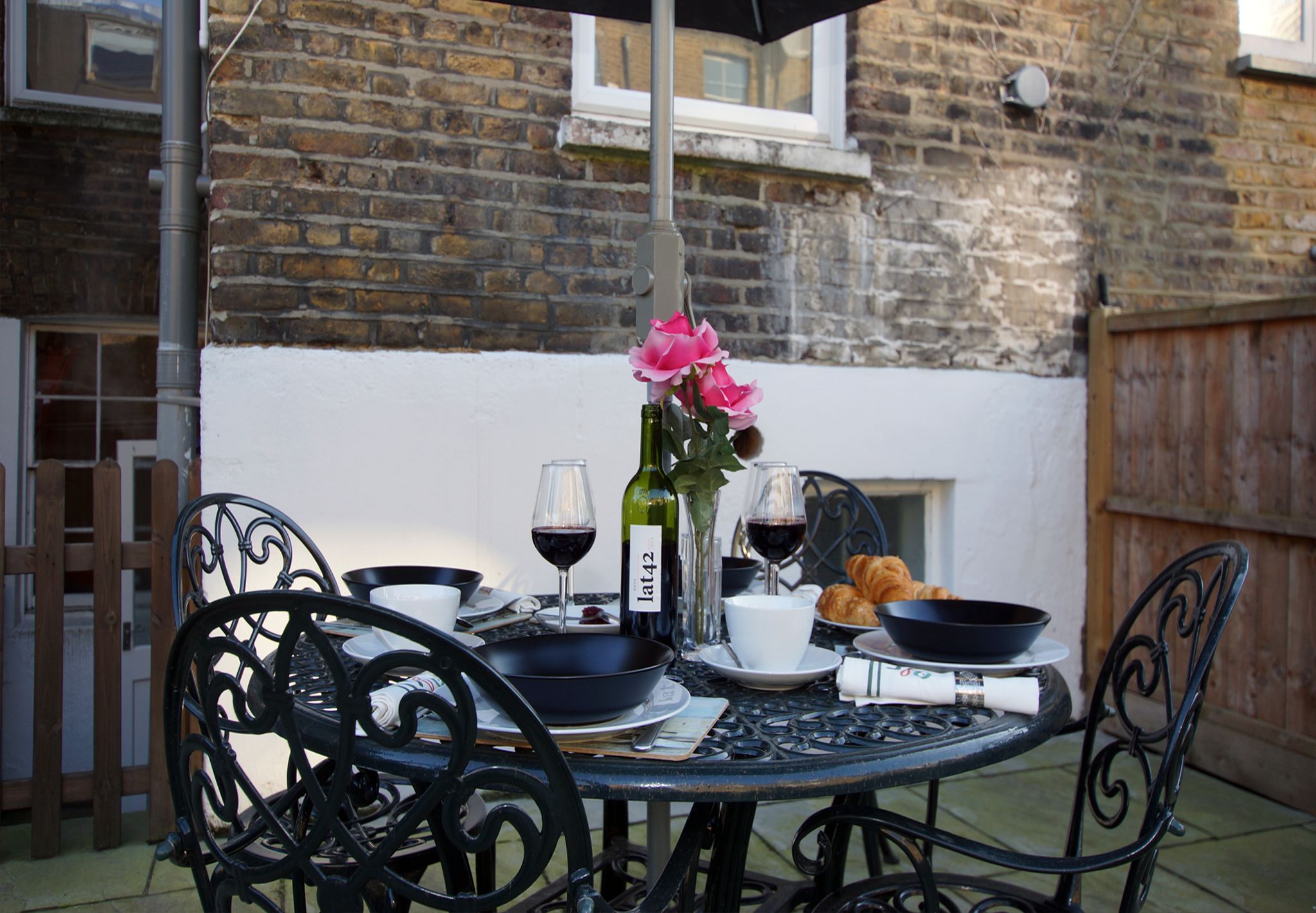 An outdoor dining table with a bottle of wine, glasses, a bowl, and a croissant.