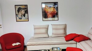A living room with a sofa, two striped pillows, a red armchair with an open book, and three framed artworks on the wall.