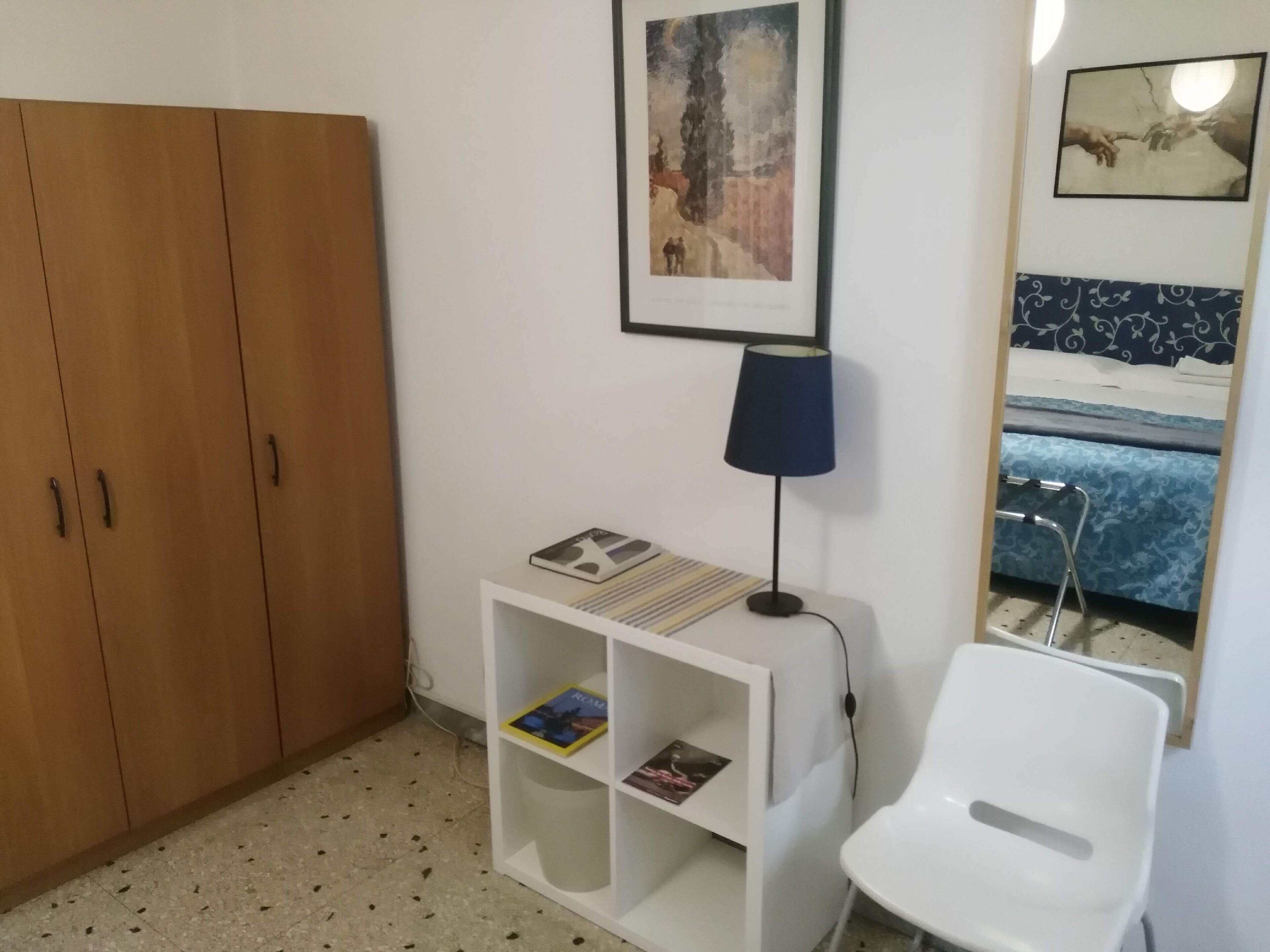 A room with a wooden wardrobe, a white shelf with books, a desk lamp, a white chair, and a bed with a blue patterned bedspread.