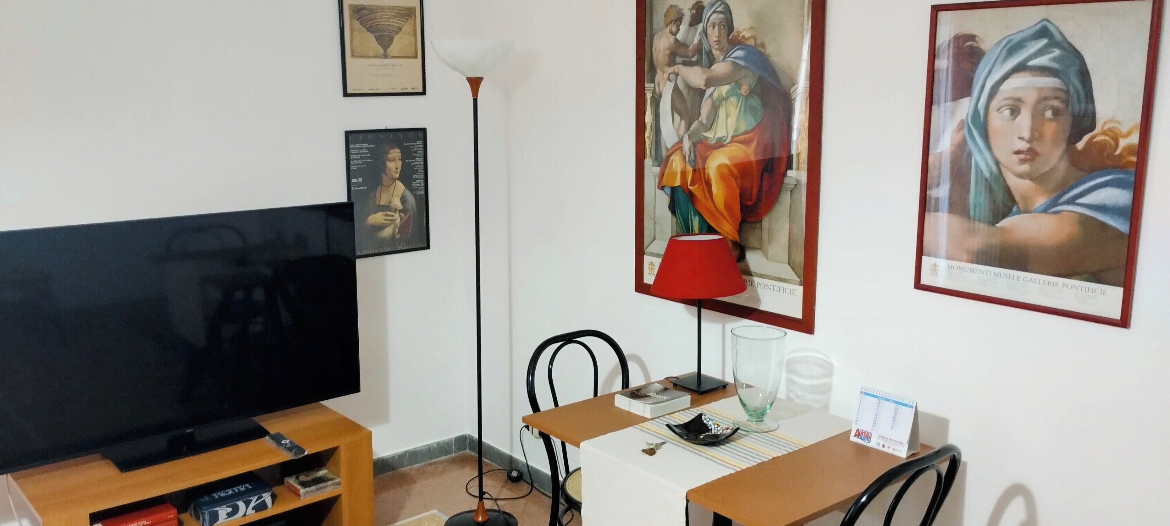 A living room with a flat-screen TV, a red floor lamp, a small table with a glass, and framed artwork on the wall.