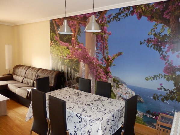 Un coin repas avec une table dressée pour quatre personnes, un canapé et une fresque représentant un paysage côtier.