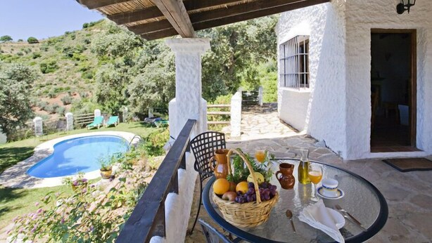 A covered outdoor area with a pool, a table with fruit and drinks, and a white building with a window.
