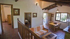 A spacious living area with a wooden staircase, a sofa, a dining table with chairs, and a view of the outdoors through a window.