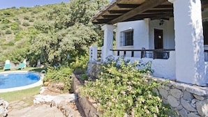 A white villa with a stone foundation, a wooden roof, and a pool.