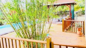 Una terraza de madera con piscina, rodeada de abundante follaje y un pabellón.