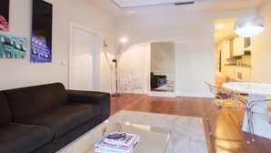 A modern living room with a dark sofa, a glass coffee table, and a dining area with a white table and clear chairs. - Top Design Apartment at OPERA Metro station (Royal Theater) / HIGH SPEED WiFi (Madrid)