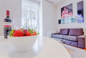A modern living room with a white table, a bowl of strawberries, two wine glasses, and a bottle of wine. There is a grey sofa and artwork on the wall. - Top Design Apartment at OPERA Metro station (Royal Theater) / HIGH SPEED WiFi (Madrid)