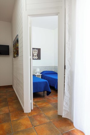 Two beds are available in this cozy room, each with blue bedding. A small table and lamp are nearby, and a framed picture adorns the wall.