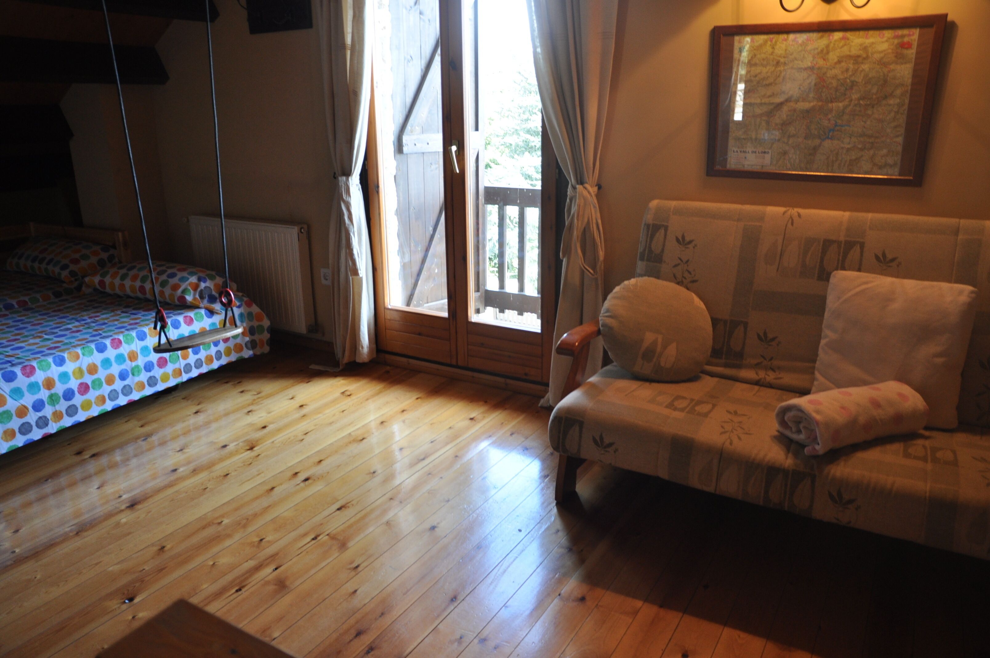 Una habitación con suelo de madera, una cama con colcha de lunares, un sofá con funda estampada y una ventana con cortinas.