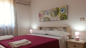 Un lit bien fait, avec une couvre-lit rouge et des oreillers blancs, deux lampes de chevet, un climatiseur fixé au mur et un tableau floral au-dessus du lit.