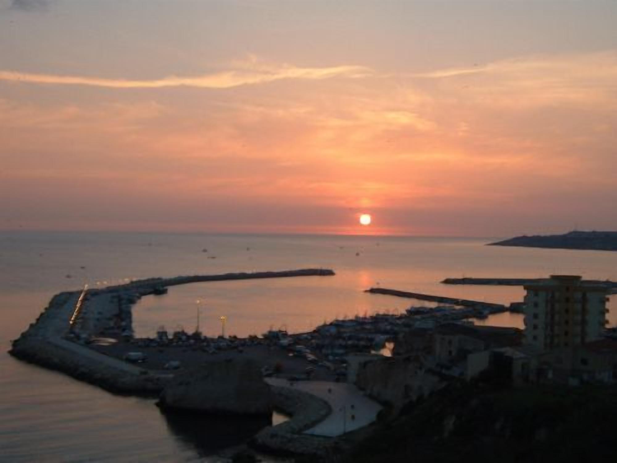 A coastal town at sunset with a calm sea, a pier, and buildings along the waterfront.