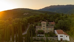 Un grand bâtiment en pierre à deux étages, doté d’une tour centrale, entouré d’une végétation luxuriante et avec des collines en arrière-plan.