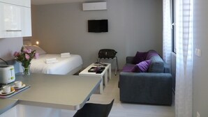 Quarto de hotel moderno com cama, sofá, pequena mesa de jantar com chávenas e torradeira, televisão na parede e janela com cortinas.