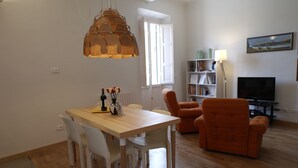 Una sala de estar moderna con una mesa de comedor de madera, sillas blancas, un sillón naranja, un televisor y una estantería.