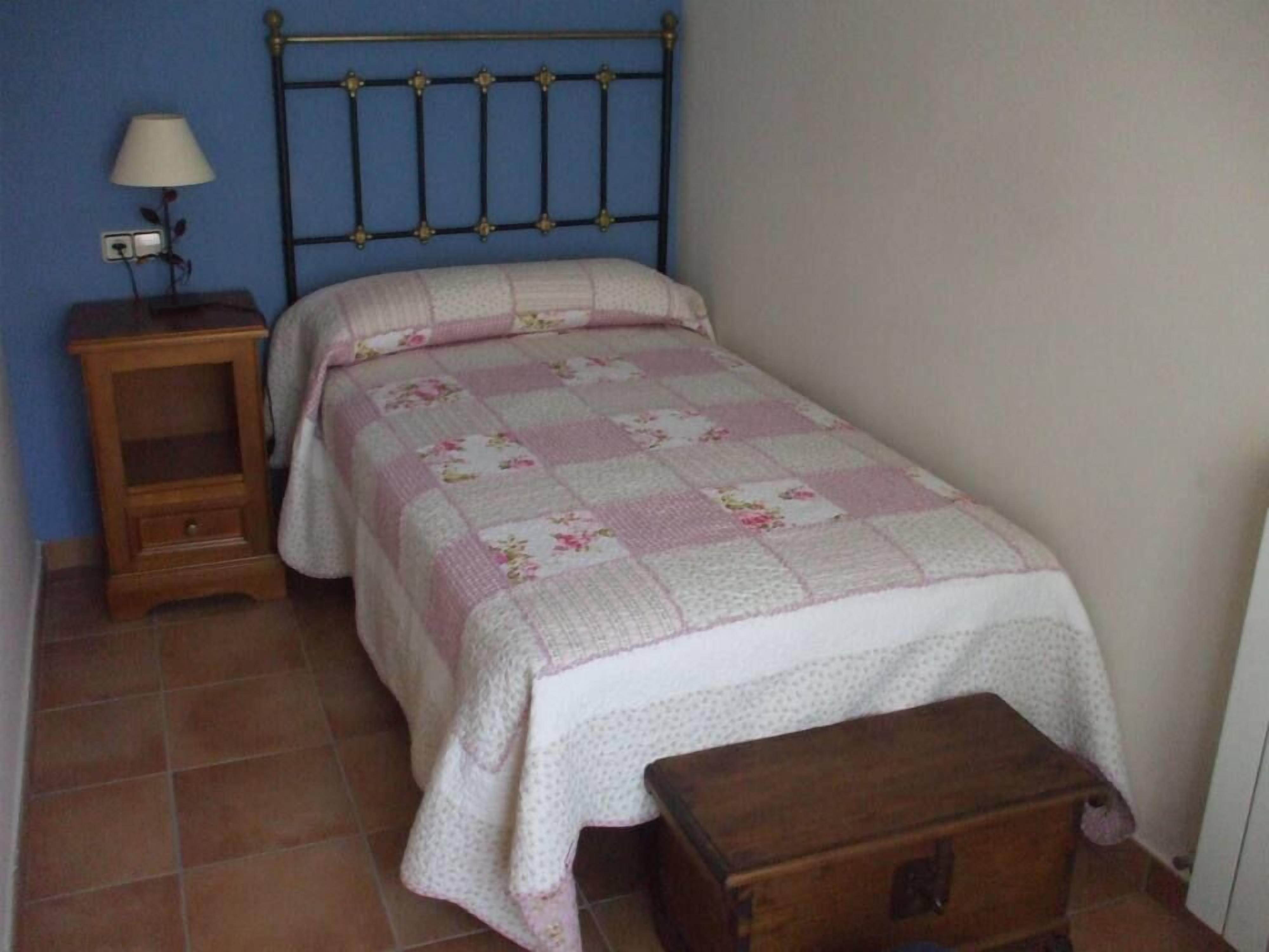 A small, single-bed room with a blue headboard, a wooden nightstand with a lamp, and a small wooden chest.