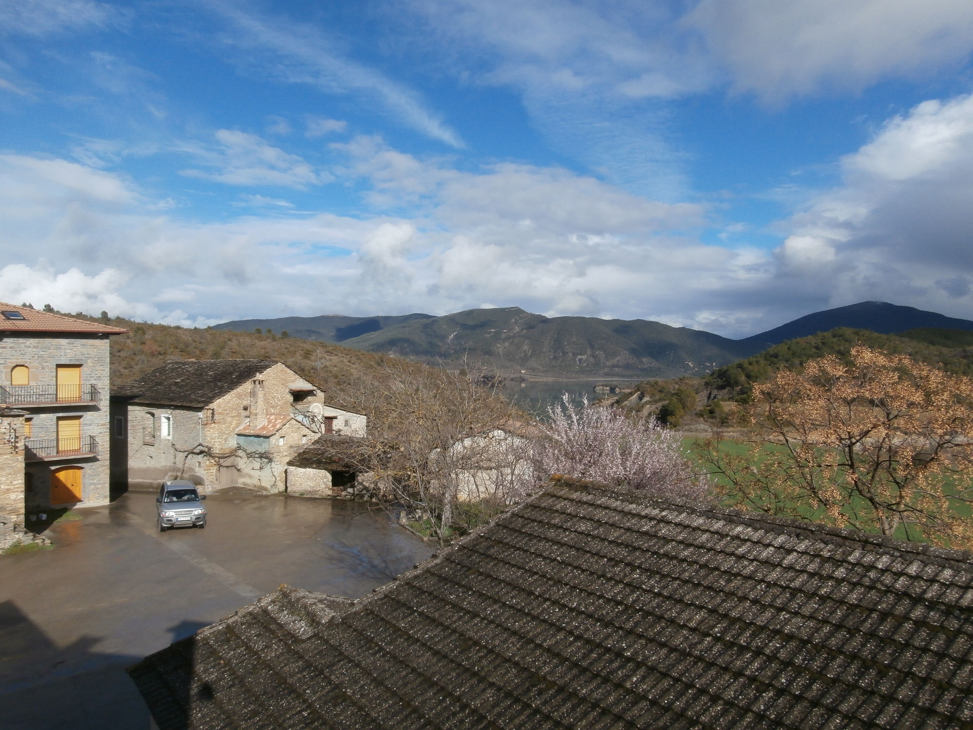 A traditional stone building with a car parked in front, surrounded by a mountainous landscape and a clear sky.