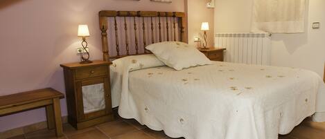 Un dormitorio con cabecera de madera, dos mesitas de noche con lámparas, cama con colcha blanca y un radiador.