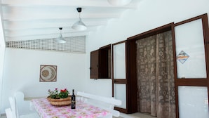 Une pièce avec une table blanche recouverte d’une nappe à motifs floraux, une bouteille et des verres, un panier en osier rempli de fleurs et un élément décoratif fixé au mur.