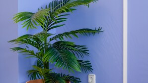 Una habitación con una planta grande en maceta, una jaula blanca con un pajarito y un cuadro en una mesita.