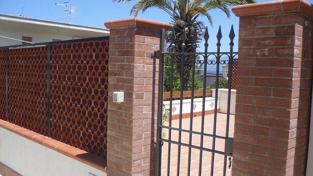 A gated entrance with a brick wall and metal gate, a palm tree, and a clear blue sky. - NEAR THE SEA AND AWAY FROM NOISES (alcamo marina)