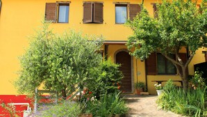A yellow building with a central entrance, surrounded by lush greenery and potted plants. - 5-bedroom holiday apartment: nature, swimming pool, relax 30 mins from the beach (Pesaro-Urbino)